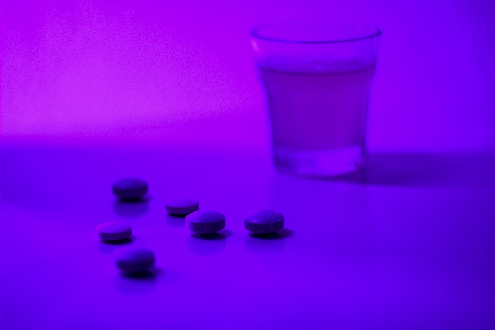 Violetilla valolla valaistulla alustalla on shottilasi ja muutama pilleri, jotka kuvaavat alkoholin ja huumausaineiden sekakäyttöä.