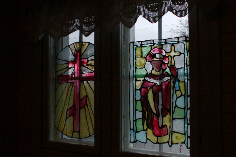 Kuvassa näkyvät majatalon ikkunat sisältäpäin. Ikkunoiden eteen on asetettu värikkäät lasimaalaukset, joista toisessa on ortodoksinen risti ja toisessa piispan hahmo.
